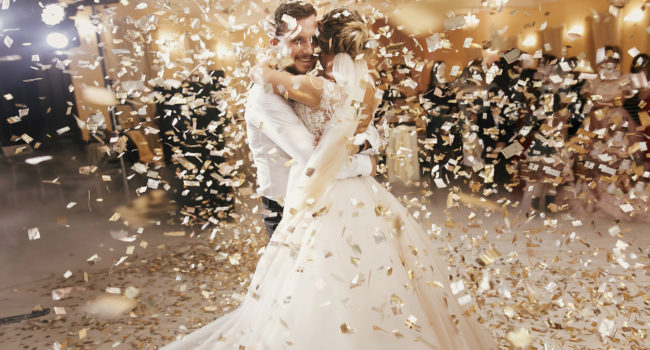 Gorgeous bride and stylish groom dancing under golden confetti at wedding reception. Happy wedding couple performing first dance in restaurant. Romantic moments