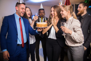 Young cheerful company workers celebrating birthday of colleague with cake and champagne in office
